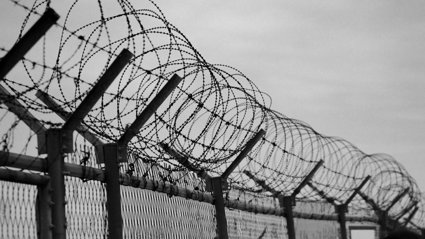 View of the top of a barbed wired fence in front of a dark cloudy sky. The image is in black and white.