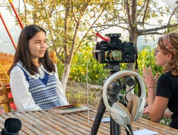 An interviewer speaks with a guest outdoors while a camera films.