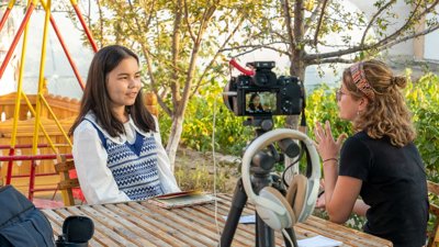 An interviewer speaks with a guest outdoors while a camera films.