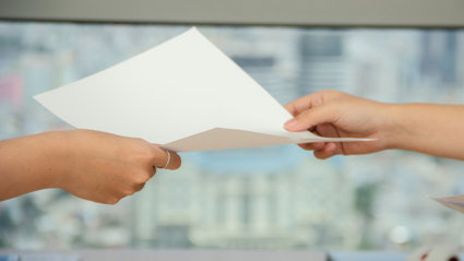 Two people hold a sheet of blank paper as one passes it to the other. Only their hands can be seen and the background is blurred.
