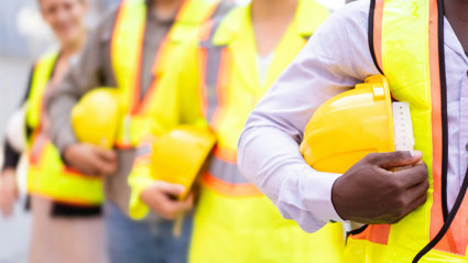 Chest-level close up of people lined up while holding hard constructions helmets and wearing yellow high vis clothing.