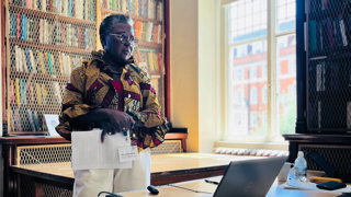 A person stands next to a table and laptop delivering a talk