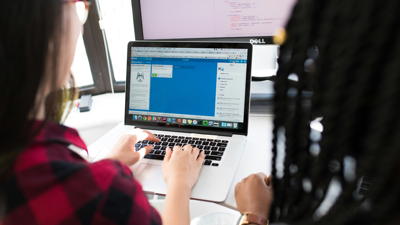 Two people sit by a laptop as one types. A desktop monitor screen sits on the table behind the laptop.