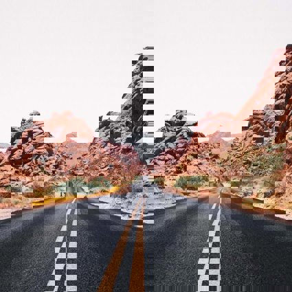 A Nevada road stretching out into the distance, the yellow central lines weaving slightly where the road bends. The road has rocky outcrops either side and in the distance