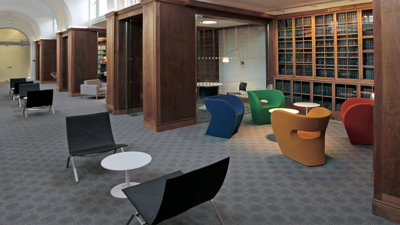 The Society's members room displaying wooden bookshelves stacked with books, plush chairs in blue, green, yellow and red. The carpet is two tones of grey and the arched celling is white.