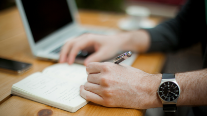 A person uses a pen pen to write on a notpad. Their phone and laptop sit next to the notepad on the wooden desk
