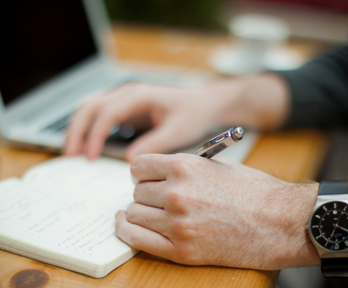 A person uses a pen pen to write on a notpad. Their phone and laptop sit next to the notepad on the wooden desk 