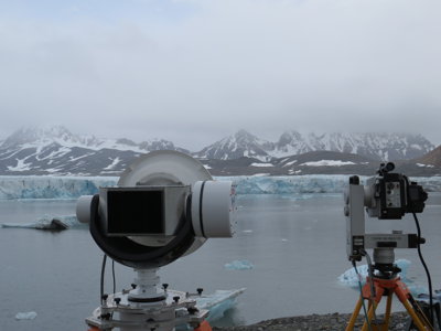 Two cameras set up on tripods pointing towards a glacier and lake. The mountains in the background are partially covered by snow. 