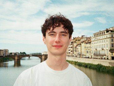 Person wearing a white t-shirt standing in front of a river with historic buildings and a stone bridge under a partly cloudy sky.