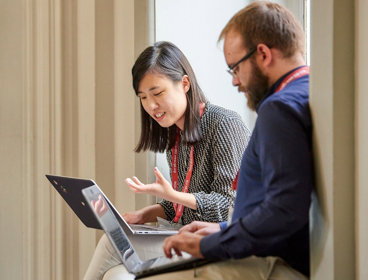 Two people sat next to each other engaged in conversation, each with a laptop on their lap.