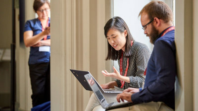 Two people sat next to each other engaged in conversation, each with a laptop on their lap.