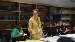Dr Alice Oates gives a talk in a library. She is pointing at various maps spread out on a table as the audience listens attentively.