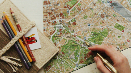 A person uses a pen to point to a location on a map. a notebook and pencils also sit on the map