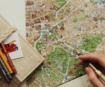 A person uses a pen to point to a location on a map. a notebook and pencils also sit on the map