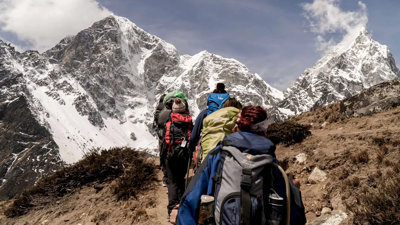 A group of people walk up mountainous terrain. They each carry large bags and walking equipment.