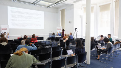 Several people sit in a modern, well-lit presentation room listening to a presenter talk.