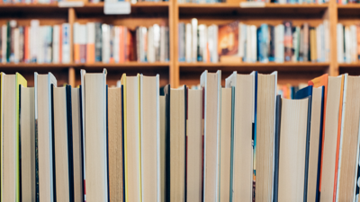 A row of books are lined up in the foreground. Behind them are blurred books shelfs full of books.