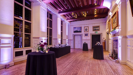 Poseur tables with floral arrangements and candles set-up for a drinks reception in a grand main hall. 