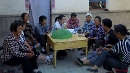 Group of people sat outside around a table. Some of them are holding pieces of paper and conversing. 