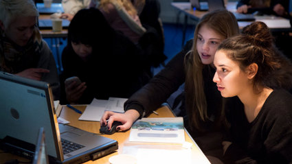 Two young students sat at a desk looking at a laptop, with one of them holding the computer mouse.