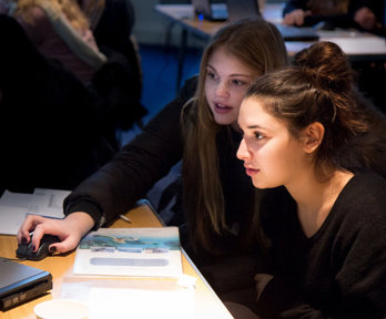 Two young students sat at a desk looking at a laptop, with one of them holding the computer mouse.