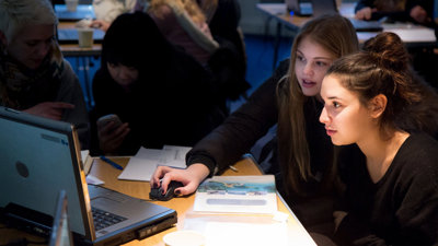 Two young students sat at a desk looking at a laptop, with one of them holding the computer mouse.