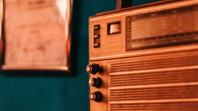 A brown retro radio in front of a dark blue wall. A blurred out framed image hangs on the wall behind the radio on the left hand side.
