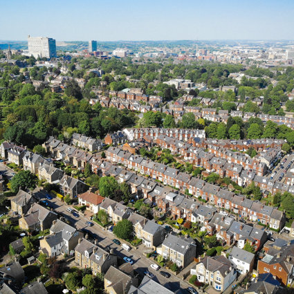 An aerial view of a residential area, with orange houses with grey roofs and small green gardens. The suburban area stretches off into the distance, with a few high rise tower blocks also in the view.