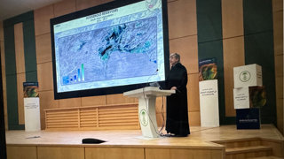 A woman on a podium giving a speech at a conference with behidn her a board that shows a map and the title Household Reservoirs South West Afghanistan from 2016 until 2019.