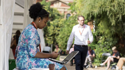 A person in a blue floral dress sits in the Society's garden with a laptop on their lap as other people sit and walk nearby.