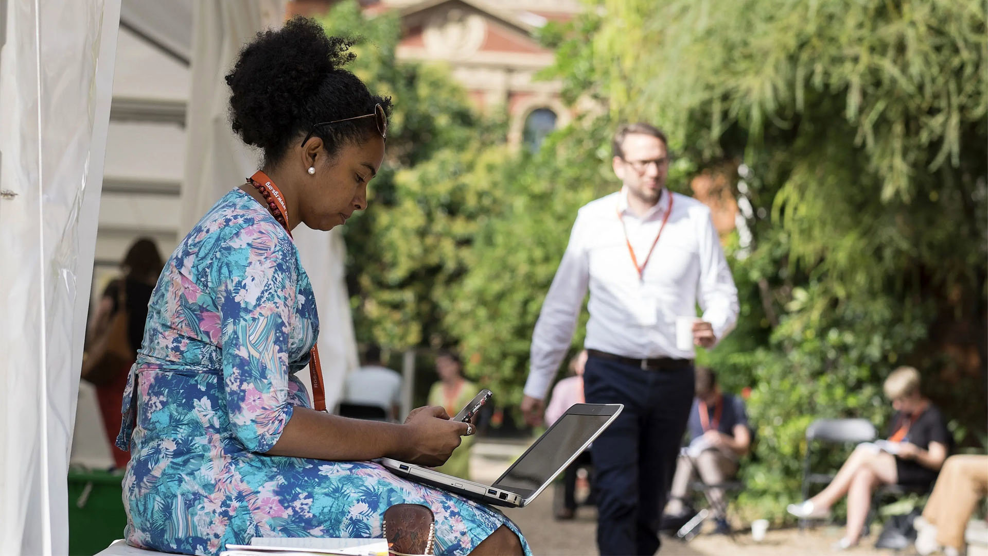 A person in a blue floral dress sits in the Society's garden with a laptop on their lap as other people sit and walk nearby.