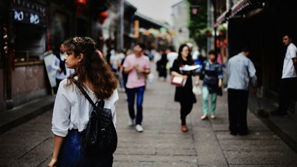 People walking on a busy street, with one person in focus as seen from behind, looking at something to their left.
