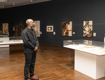 Man standing in an exhibition space, surrounded by display cases and hanging pieces of art