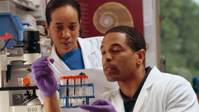 One researcher in a lab coat and disposable gloves holds a test tube rack whilst placing a test tube inside. A second researcher stands beside the first watching. 