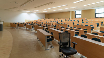 Empty lecture hall the lights on and a single office chair on the opposite side of the hall from the the lecture podium.