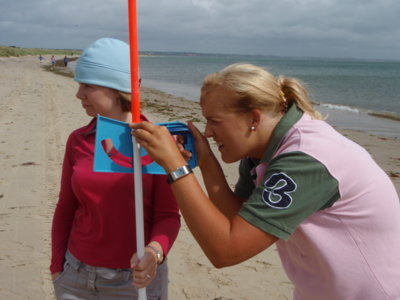 Two female students on a sandy beach, one is holding an ranging pole and the other is using an instrument to measure the height of dunes in the distance