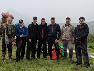 Seven people stood in a row carrying expedition equipment, with a mountain in the background.