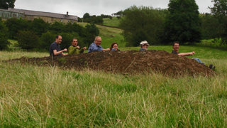 Group of people sitting in a field of grass on a elongated hill pretending to row a boat.