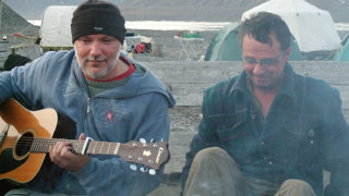 Two people in nature sitting at a campfire while one of them plays accoustic guitar.