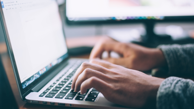 A person types on their laptop whilst sitting at a desk. a computer monitor can be seen in the blurred background. 