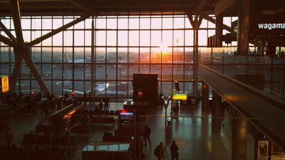 The sun rise casts light on Heathrow airport tarmac. Planes can be seen stationed on the tarmac through a large window. The dimly lit interior of the airport can be seen before the window, in the foregroun.