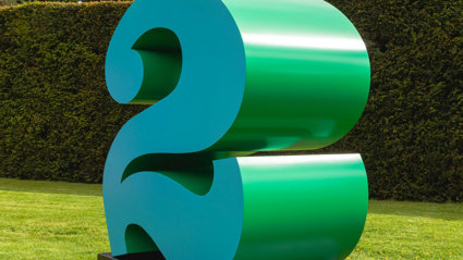 A large green number 2 on a background of grass, with a hedge and cloudy blue skies.