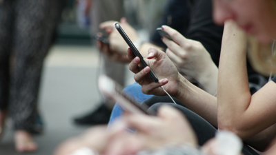 Several people sit next to each other on a public bench which is out of view. They all have mobile devices in their hands which they are each staring at.