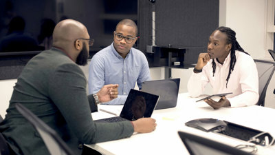 Three people sit at a desk with laptops and a tablet. One sits at the head of the table and the other two opposite each other.