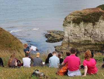 Young people sitting on a grassy sloped hill and listen to a teacher talking about the natural landscape