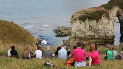 Young people sitting on a grassy sloped hill and listen to a teacher talking about the natural landscape
