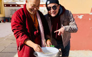 Two men smiling while preparing food outside. The person on the left wears a maroon monk robe and uses chopsticks to stir food. The person on the right points at the bowl playfully.
