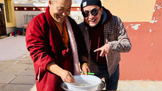 Two men smiling while preparing food outside. The person on the left wears a maroon monk robe and uses chopsticks to stir food. The person on the right points at the bowl playfully.