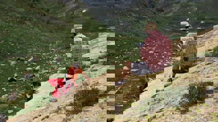 A researcher sitting on a rocky slope in a valley writing in a notebook while two others stand testing the rock hardness.