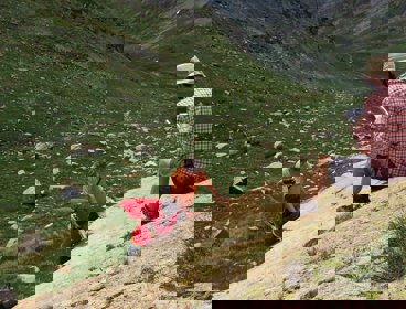 A researcher sitting on a rocky slope in a valley writing in a notebook while two others stand testing the rock hardness.